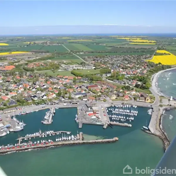 En kystby set fra luften med både i havnen, grønne marker og gule rapsmarker i baggrunden under en klar blå himmel.