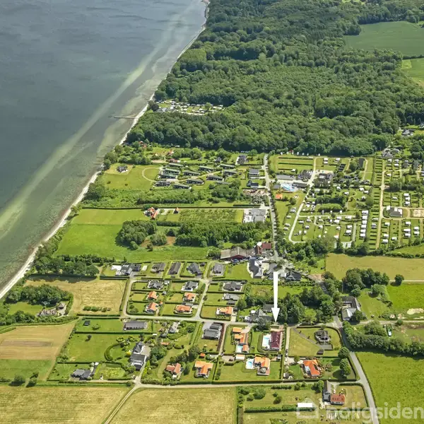 Kystlinje med roligt vand grænser op til en by med spredte huse og grønne marker. Området er omgivet af træer og landbrugsjord, med en campingplads i det fjerne.