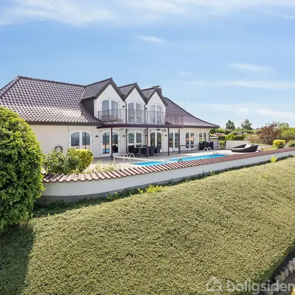 En stor, hvid villa med mørkt tag står i en velplejet have. Foran huset er en swimmingpool omgivet af terrasseområde. Solrigt vejr og få skyer på himlen.