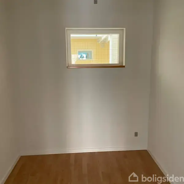 Et lille rum med hvide vægge og trægulv. Et lille vindue øverst på væggen viser udsigt til en gul murstensbygning udenfor. Ingen tekst synlig.