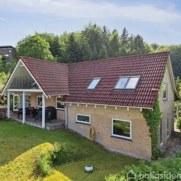 Huset med rød tag og murstensvægge står i et grønt område. En overdækket terrasse med siddepladser ses ved siden af huset. Skov omkranser ejendommen.