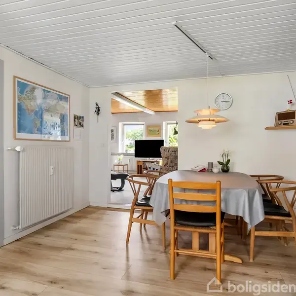 Et spisebord med seks stole er dækket af en grå dug. Bordet står i et lyst rum med træloft og trægulv. I baggrunden er et opholdsområde med computer og stol.
