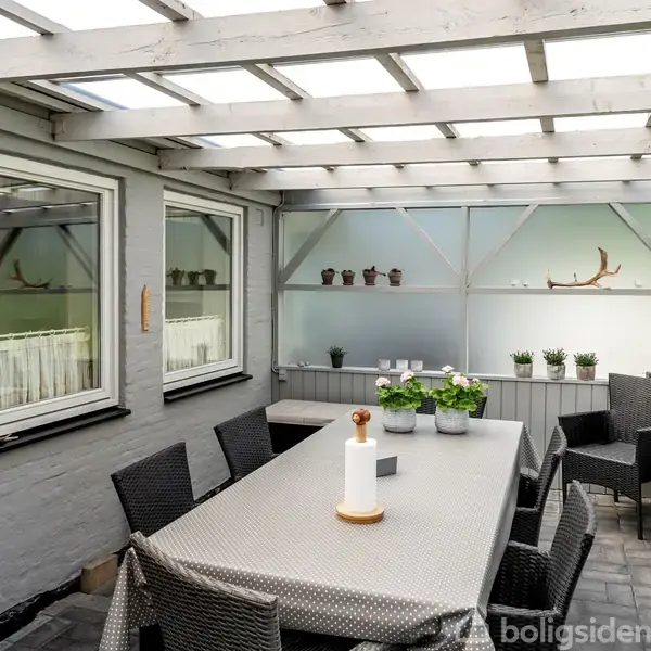 Langt bord med sort-flettede stole står under overdækket terrasse. Bordpynt inkluderer planter og lysestage. Mure og vinduer omgiver terrassen, som har et lyst tag og grønne planter på hylder.