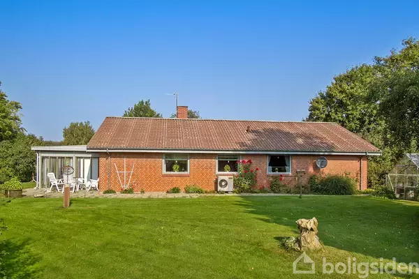 Rødstenshus med fladt tag står midt i en frodig have. Der er en terrasse med havemøbler til venstre og omgivet af træer mod en klar blå himmel.