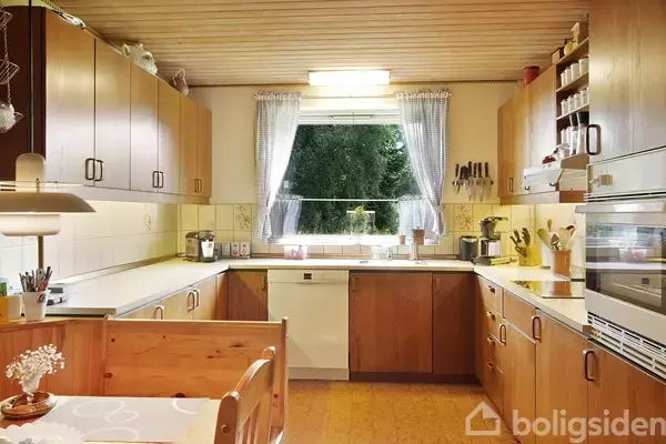 Et lyst køkken med træskabe er fyldt med køkkenredskaber. Sollys strømmer ind gennem et vindue med ternede gardiner. Bordet har en vase med blomster.