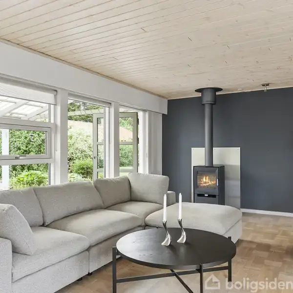 Sofagruppe i moderne stue. Sofa står ved store vinduer med udsigt til have. Pejs i hjørnet, sort væg bagved, og rund bord med stearinlys.