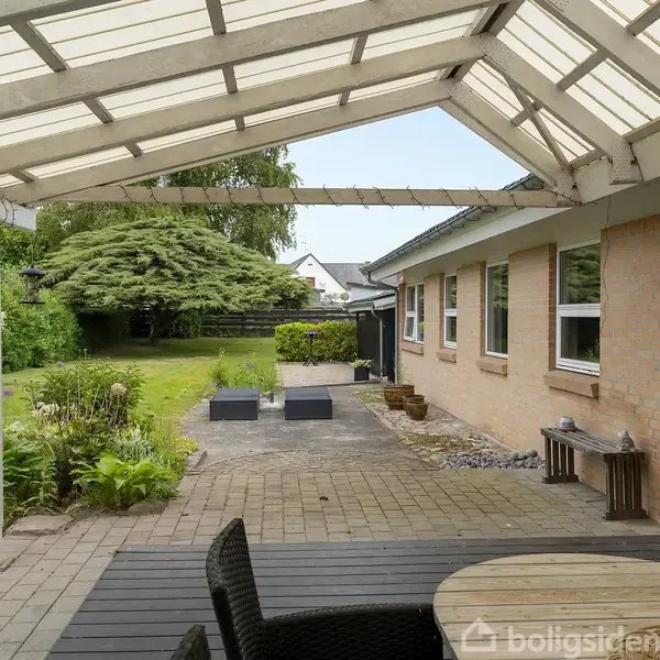En overdækket terrasse med bord og stole står ved et hus af mursten. I baggrunden ses en grøn have med buske og træer.