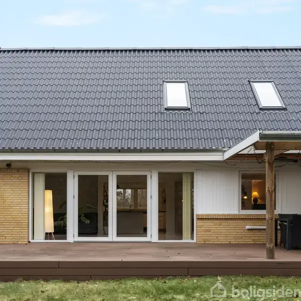 Hus med mørkt tag og gul murstensfacade, der står stille i en have med en træterrasse og overdækket siddeområde. Vinduer og døre åbner ud til terrassen.