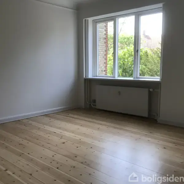 Et lyst rum med trægulv og et stort vindue. Vinduet vender mod en grøn have og lukker naturligt lys ind. Væggene er hvide og radiatoren er under vinduet.