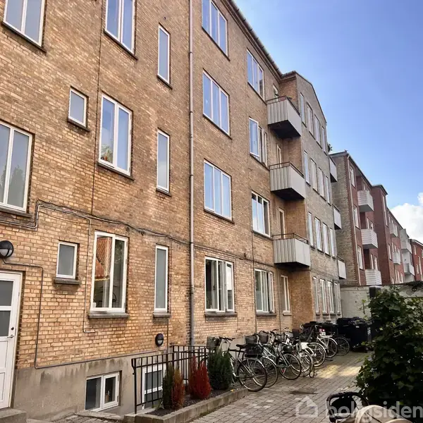 En murstensbygning viser flere etager med vinduer og små altaner. Cykler er parkeret langs facaden. Bygningen er omkranset af en gårdhave med buske under en blå himmel.