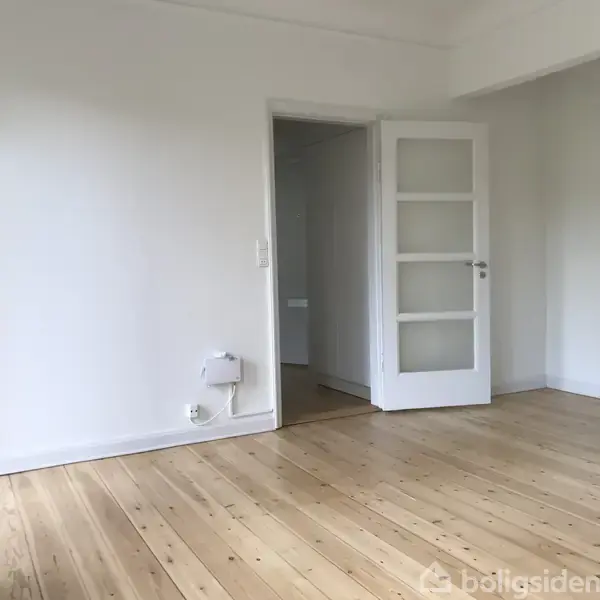 Et tomt rum med en åben hvid dør. Det har lyse trægulve, hvide vægge, en dør fører til en anden del af boligen. Ingen tekst er til stede.
