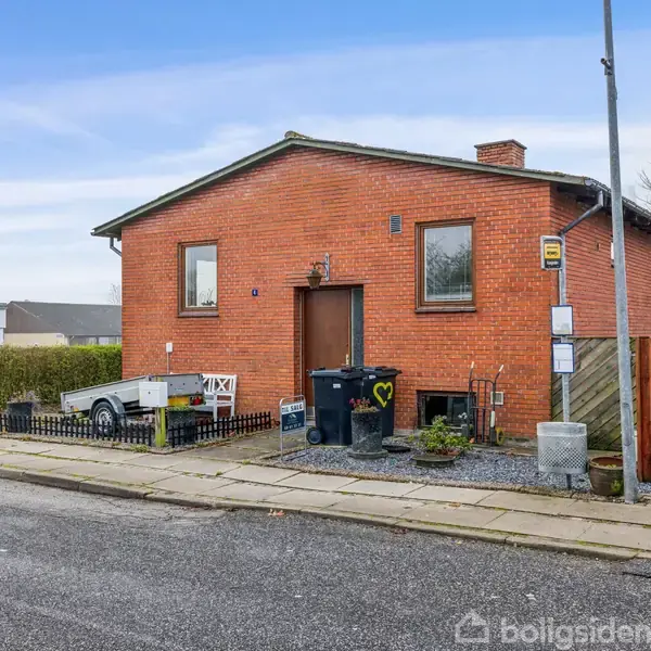 Rødt murstenshus har to store vinduer; placeret ved fortov i en villavej med en trailer parket ved siden af, samt skraldespande og en busstoppested til højre.