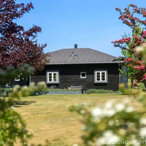 Et sort træhus står stille i en frodig have med blomstrende træer og en græsplæne under en klar blå himmel.