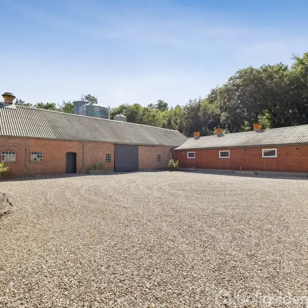 To brune murstensbygninger står overfor hinanden. Bygningerne omgiver en gårdsplads med grus, og der er tæt skov i baggrunden under en klar blå himmel.