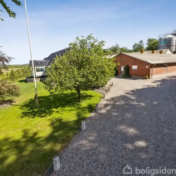 Et rødt murstenshus står ved en grusvej omringet af grønne træer og en velplejet græsplæne. En stor silo er placeret i baggrunden, nær huset.