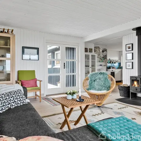 Stue med grå sofa og farverige puder, en rund stol ved et sofabord. Brændeovn opvarmer rummet. Lysindfald fra vinduer, omgivet af lyse møbler og flettede kurve.
