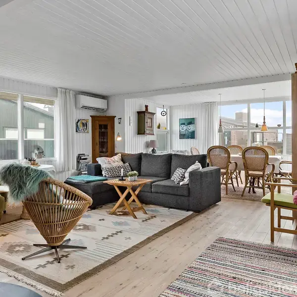 Stue med sofaarrangement og TV, omgivet af lyse trævægge og gulv. Spisebord med stole i baggrunden. Rum indrettet med tæpper, kunst på væggene, og store vinduer til venstre.