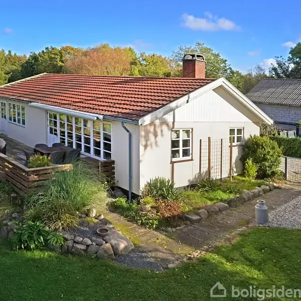 Et hvidt hus med rødt tegltag står i en have med grøn plæne. Huset har store vinduer, en terrasse omkranset af plantebede, og ligger i et landområde med træer i baggrunden.