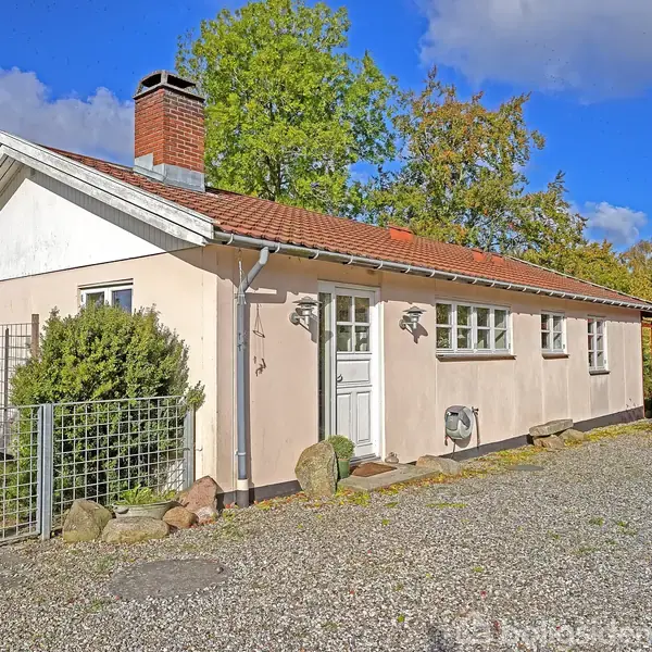 Et hvidt hus med rødt tag står stille. Foran huset er der en grusbelagt indkørsel og en metalport. Baggrunden har grønne træer og en blå himmel.
