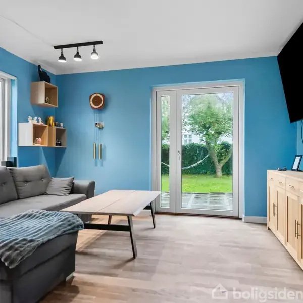 Grå sofa med tæppe vender mod rektangulært sofabord i blå stue. Store vinduer og glasdøre med udsigt til have. Væghængt TV og træskænk til højre.