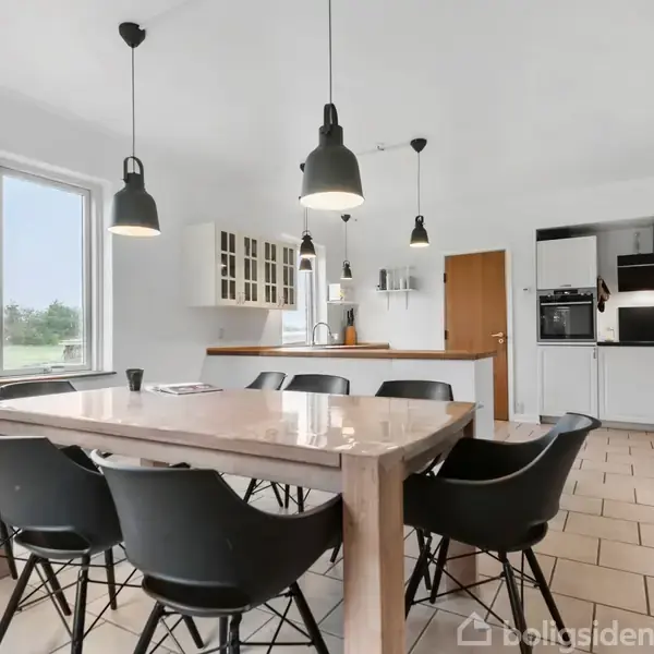 Et stort træbord med seks sorte stole står i et lyst køkken med flisegulv. Der hænger tre sorte lamper over bordet, og der er udsigt til en have gennem vinduerne.