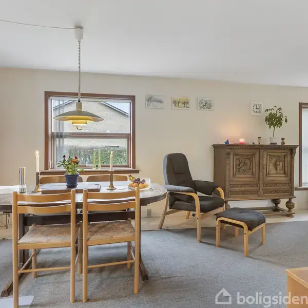 Rundt spisebord med fire stole står i en stue. Ophængt lampe over bordet. Fritstående skænk til højre, med pyntegenstande. Vinduer giver udsigt til haver udenfor.