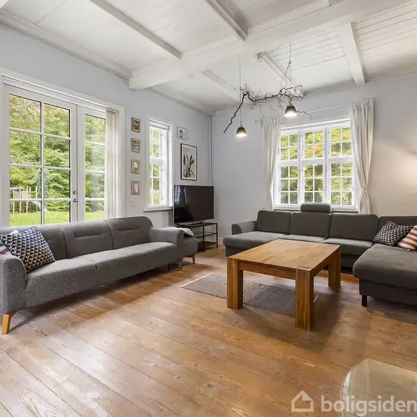 En moderne stue med to grå sofaer står i et lyst rum. Et træbord står mellem sofaerne, og store vinduer viser en grøn udsigt udenfor. En sort tv-skærm står i hjørnet.