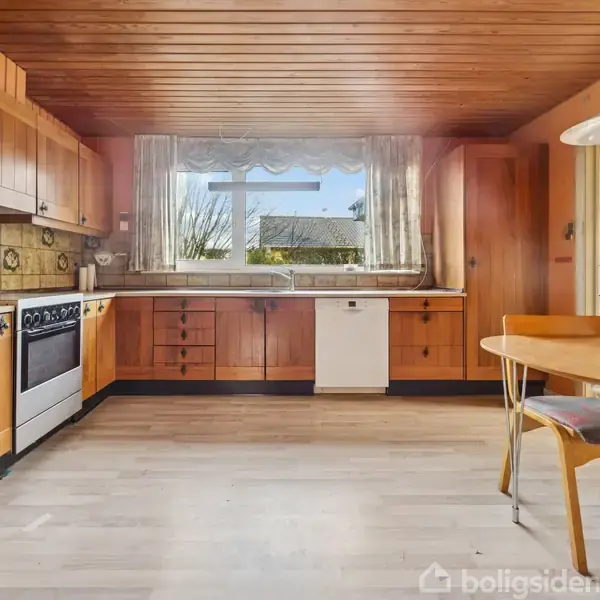 Et køkken med trækabinetter og komfur placeret ved et vindue. Hviskede gardiner hænger over vinduet, der giver udsigt til en have. Rundt bord med stole i højre side.