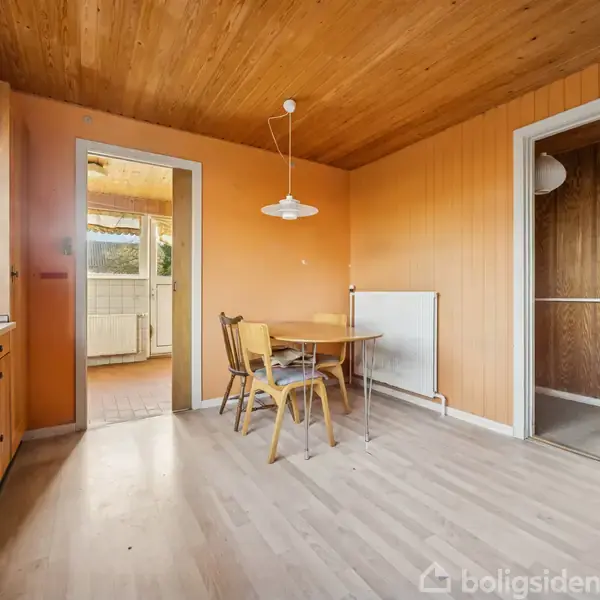 Køkkenbord med stole står i et rum med trævægge og -loft, ved siden af en radiator. To døråbninger fører til en entré og et solrigt rum. Ingen tekst fundet.