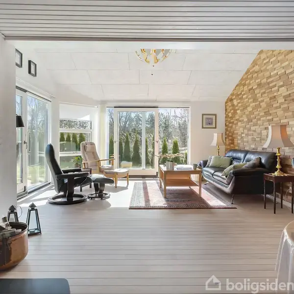 Stue med brændeovn til venstre, sofa og lænestole i midten, vinduer med udsigt til haven, og en væg med træpaneler til højre.