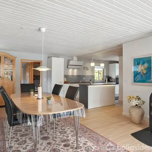 En spisestue med et ovalt træbord omgivet af sorte stole. Der er en brændeovn, der brænder. Baggrunden viser et lyst køkken med hvide skabe. En farverig maleri hænger på væggen.