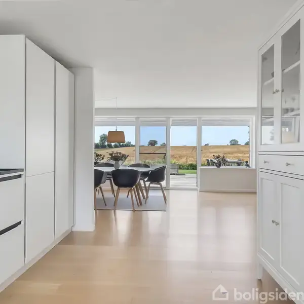 Et moderne køkken med hvide skabe og indbygget ovn fører til en spisestue med et bord og stole. Store vinduer viser en udsigt til et åbent landskab.
