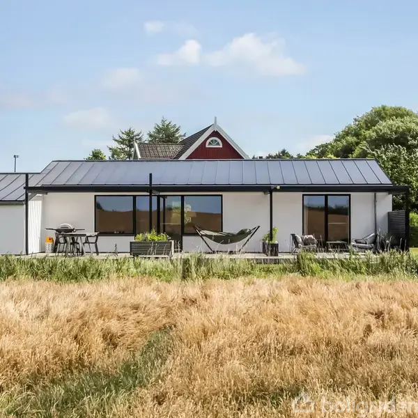 En moderne hvid villa med fladt tag står stille på en velplejet terrasse med havemøbler i en frodig, grøn have. I forgrunden er en kornmark under en blå himmel.