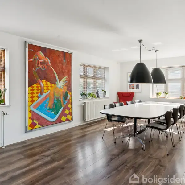 Et moderne spisestuelokale med et langt, hvidt bord omgivet af sorte stole. Over bordet hænger tre sorte lamper. Væggene har store malerier og vinduer med planter.