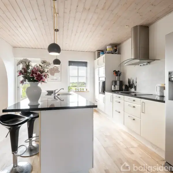 Et moderne køkken med en stor køkkenø, hvorpå der står en vase med blomster. To barstole står ved øen. Det lyse rum har hvidt skab og stål-apparater.