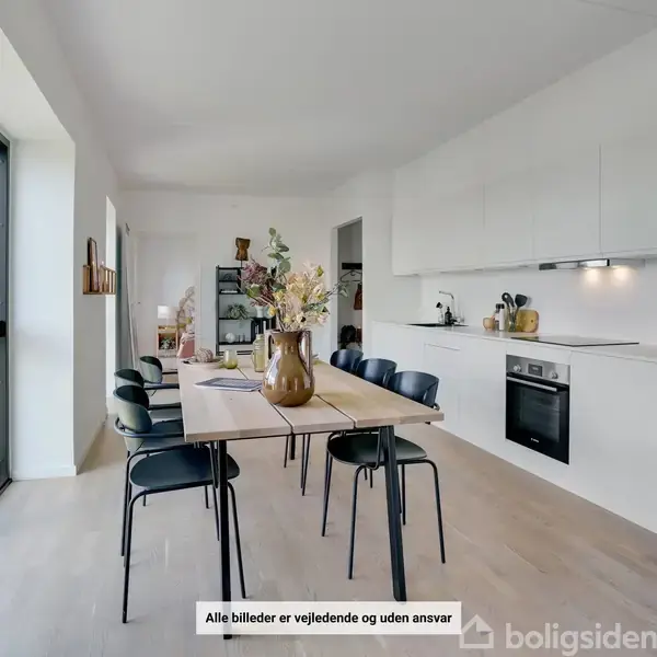 Et spisebord med seks stole står i et lyst, moderne køkken. Køkkenet har hvide skabe, indbygget ovn og dekorationer. Store vinduer giver naturligt lys. Tekst: "Alle billeder er vejledende og uden ansvar".