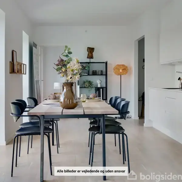 Spisebord prydet med blomster i vase, omgivet af sorte stole, står centralt i moderne, lys spisestue med åbent køkken. Tekst: "Alle billeder er vejledende og uden ansvar".