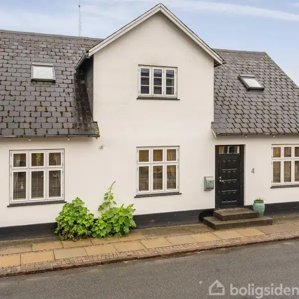 Et hvidt hus med sort tag står stille på en flisebelagt fortov i et boligområde. Nummeret "4" er synligt ved døren.