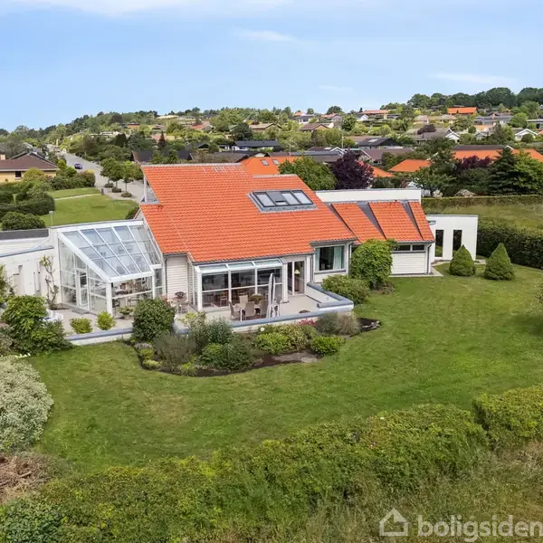 Et moderne hus med rød tegltag står omgivet af en velplejet have. Store vinduer og en udestue skaber en lys atmosfære i et kuperet villakvarter.