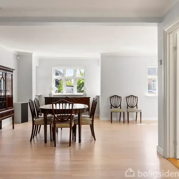 Et rundt spisebord omgivet af stole står i et lyst rum med trægulv. En stor skænk står til venstre, og flere vinduer lukker naturligt lys ind.