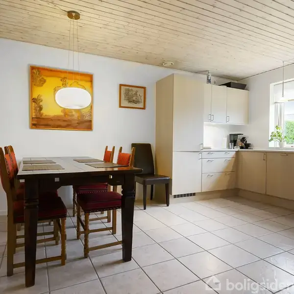 Et lyst køkken har hvide skabe og apparater som en ovn og mikroovn. To loftslamper hænger over et vindue med planter. Fliser dækker gulvet.