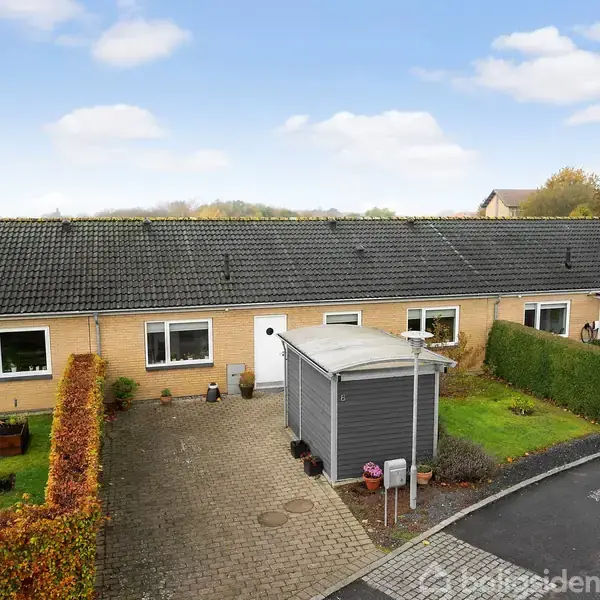 Rækkehuse står på linje, omgivet af velplejede haver og hække, i et landligt område med græsarealer og marker i baggrunden under en klar blå himmel med spredte skyer.
