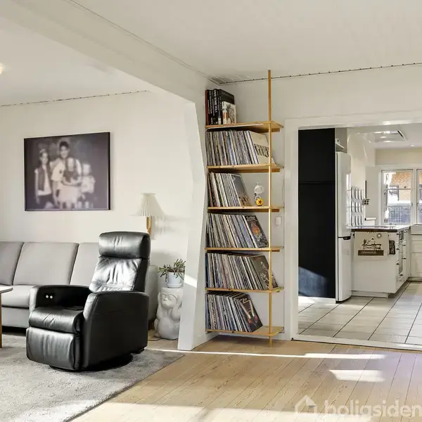 Stue med grå sofa og sort lænestol ved et sofabord. Reol med LP-plader står i hjørnet. Åben adgang til et lyst køkken med hvide skabe i baggrunden. Maleri på væggen.