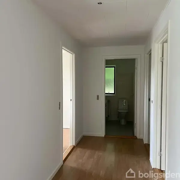 En hvidmalet gang med trælaminatgulv leder til et badeværelse. Badeværelset har et vindue og indeholder et toilet. To åbne døre ses på hver side af gangen.