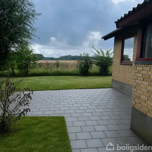 En flisebelagt terrasse ved en gul murstensbygning står rolig i forgrunden, omgivet af græsplæne og buske, med en udsigt til en mark under en overskyet himmel.