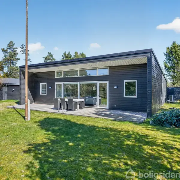 Et moderne, sort sommerhus står stille på en velholdt græsplæne. Terrassemøbler er opstillet foran store vinduer. Omgivet af træer og blå himmel i en rolig have.