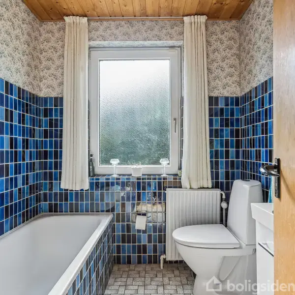 Et badeværelse med blå fliser omkranser et badekar og en toiletstol. En enkeltvindue med gardiner lukker lys ind. Gulvet er mønstret i neutrale farver.