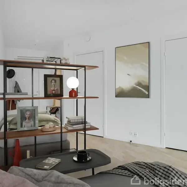 En reol med bøger, billeder og en lampe står i et lyst værelse. Væggen har en dør og et stort, abstrakt maleri, og sofaen er i forgrunden.