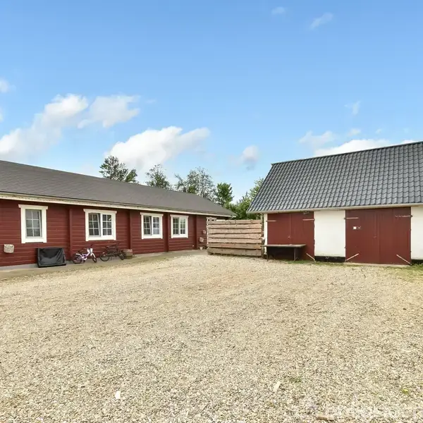 Rød bygning med hvide vinduer og cykler foran, står til venstre. Til højre ses en mindre bygning med gråt tag. De står på en grusbelægning under en blå himmel.