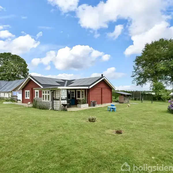 Et rødt hus med en udestue står på en stor grøn græsplæne omgivet af træer. Et lille blå bord med stole ses til højre.